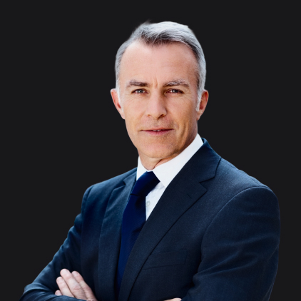 About: A middle-aged man with short gray hair, wearing a dark suit, white shirt, and navy tie, stands confidently with arms crossed against a plain black background.