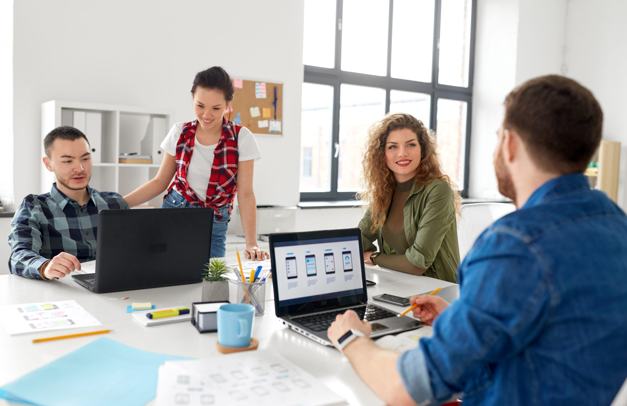 Four people sit and stand around a table in a bright office, collaborating with laptops, papers, and coffee cups. They appear to be discussing details about a project, with app designs visible on a laptop screen.