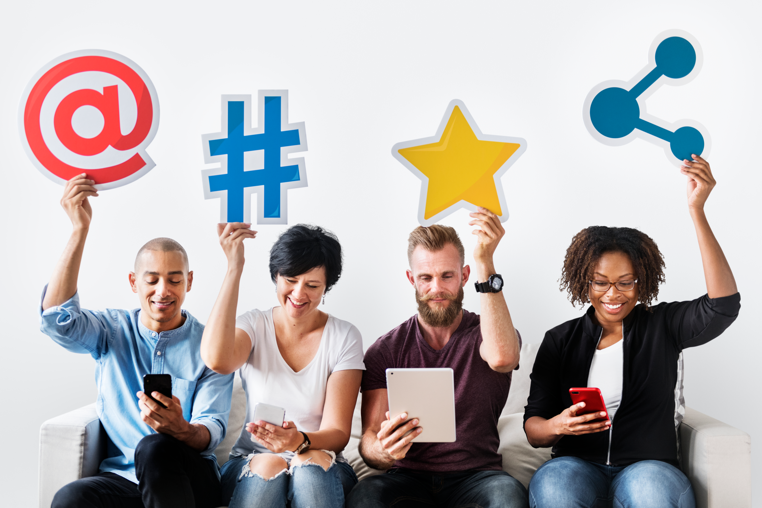 Four people sitting on a couch using digital devices, each holding up a large symbol—an at sign, a hashtag, a star, and a share icon—showcasing what social media is all about: online interaction and digital connection.