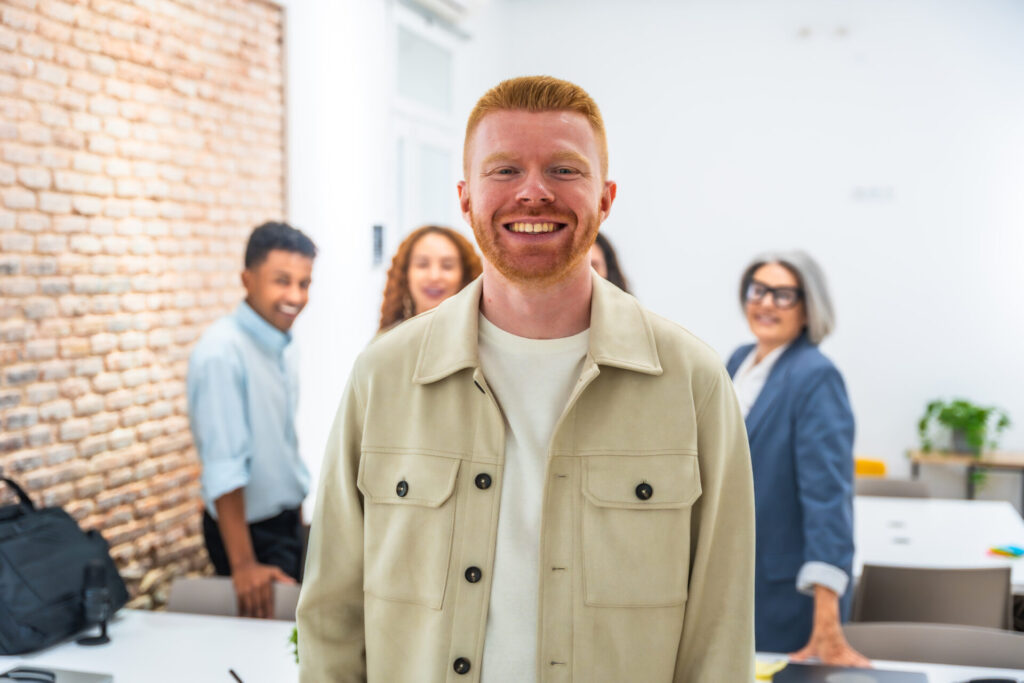 A young man with red hair and a beige jacket smiles at the camera in a modern office, ready for a social media post, with three colleagues standing and smiling in the background.