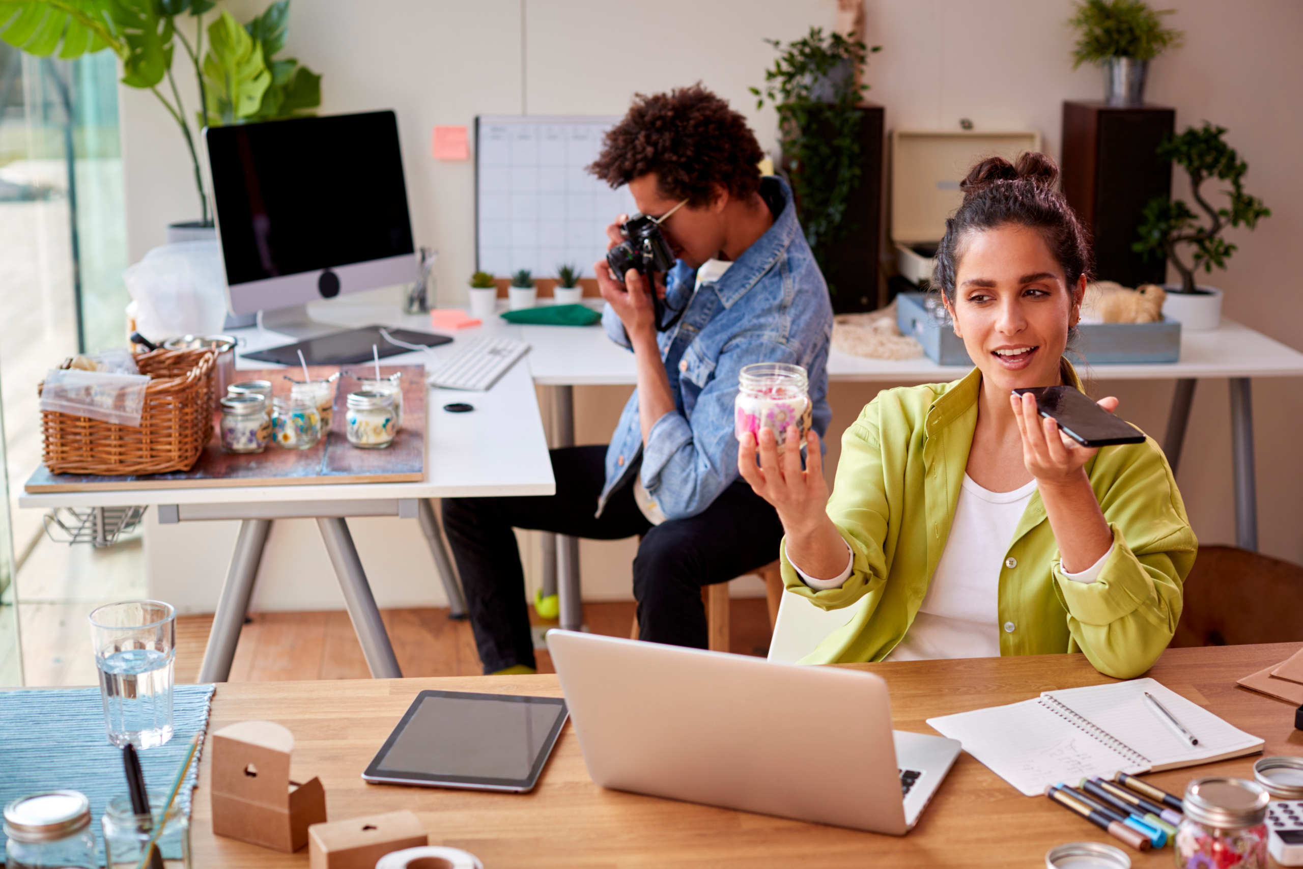 About: Two people work at a creative office desk; one woman holds a jar and speaks into her phone, while another person photographs products nearby. Laptops, jars, and plants are seen around them.